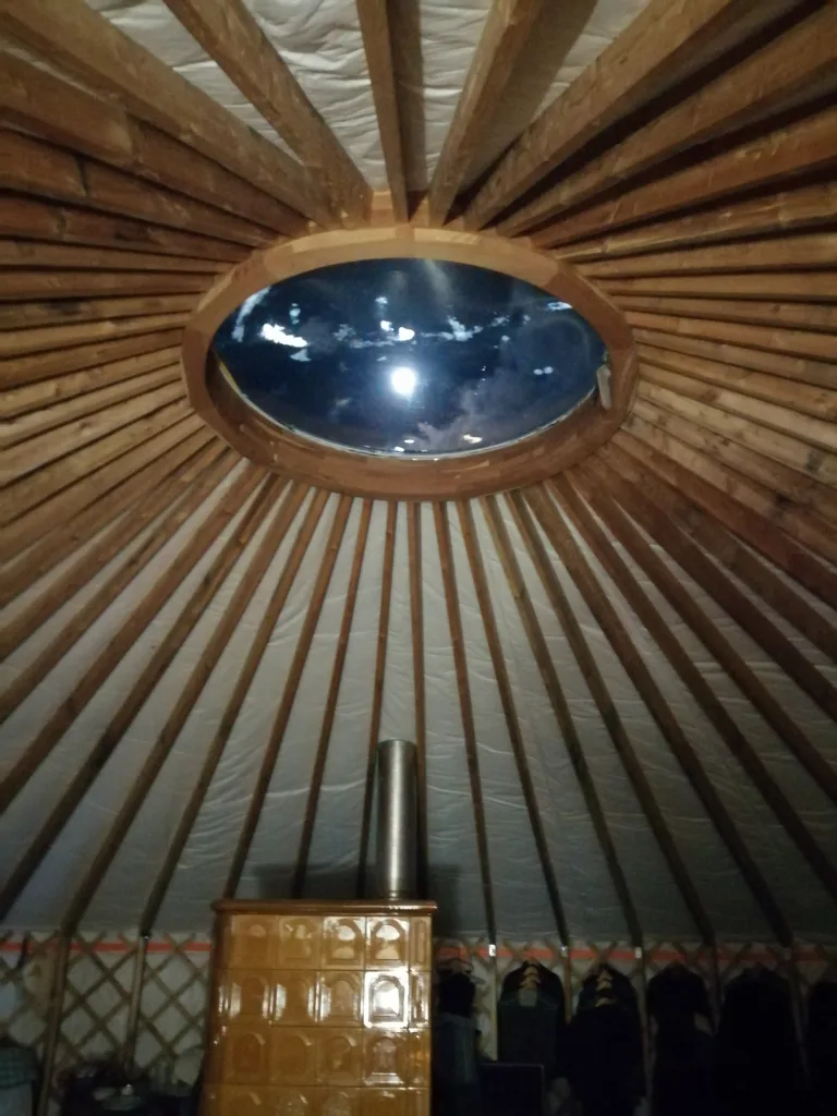 The warm interior of Barna's own yurt in the evening: the glow of the tiled stove and the roof window, golden-wood lattice wall