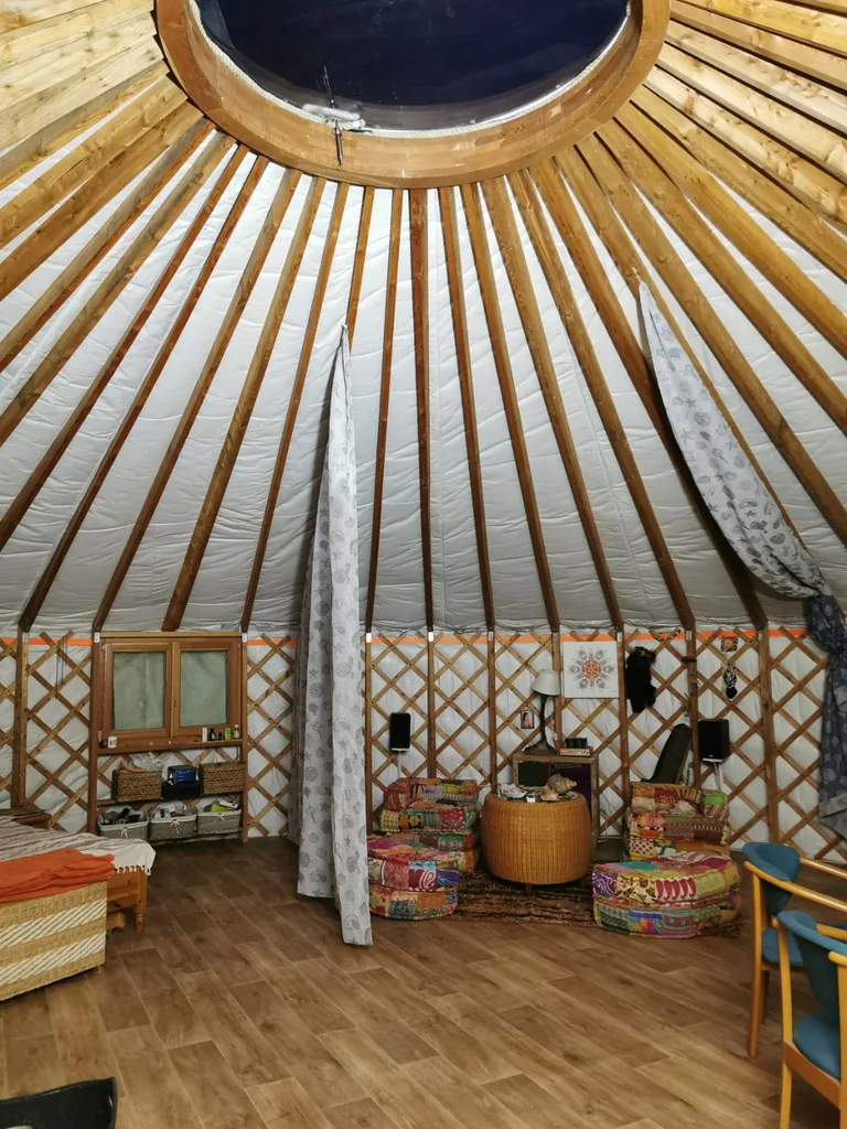 Yurt interior looking up to the wooden crown ring and roof window, with floor cushions on a kilim rug below