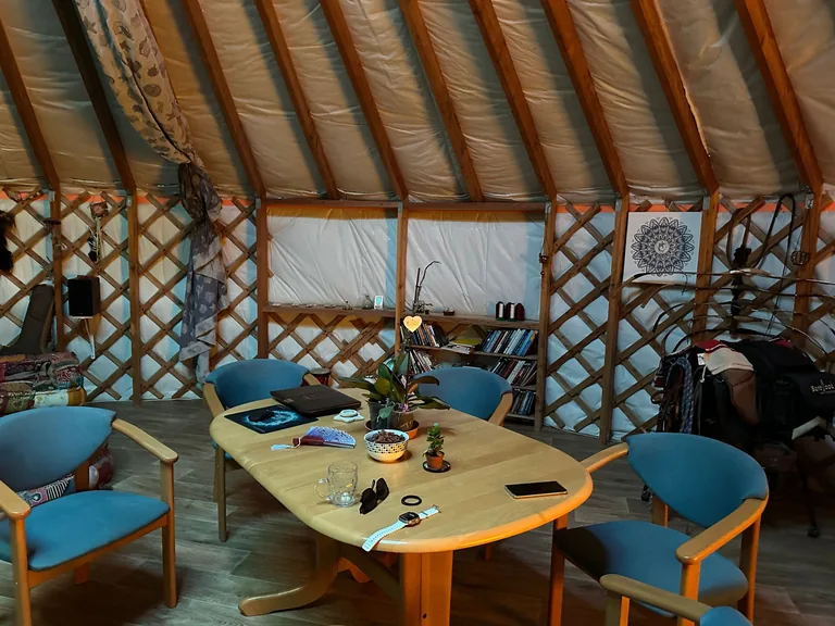 Furnished yurt interior: oval wooden table, blue armchairs, mandala wall art and a bookshelf, a homely living room