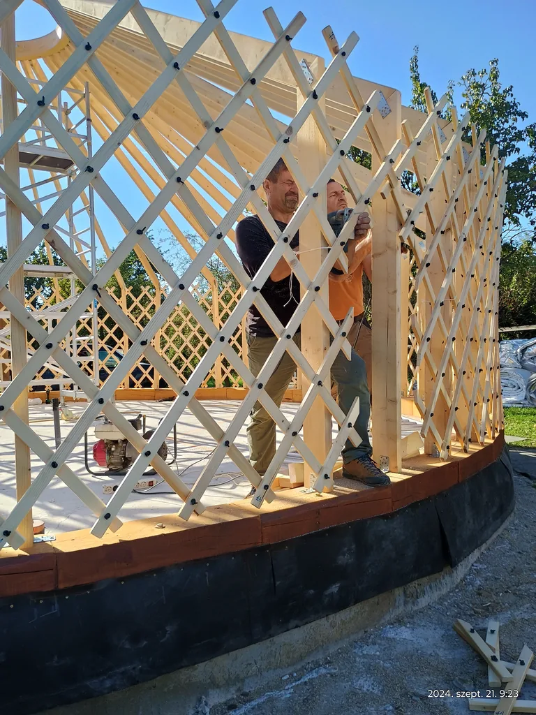 The wooden roof structure and lattice wall of the yurt during construction, two builders inside the frame, blue sky
