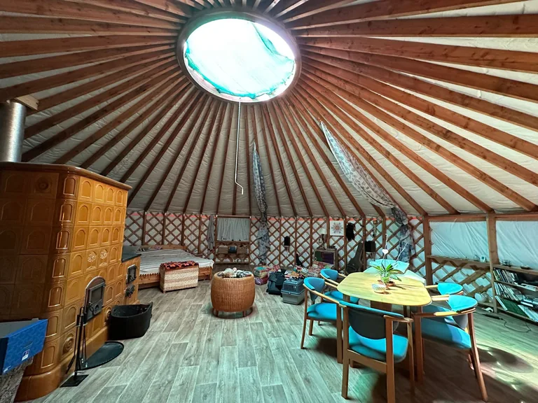 Yurt interior with a tiled stove and oval roof window, wooden table with blue chairs, a warm living space